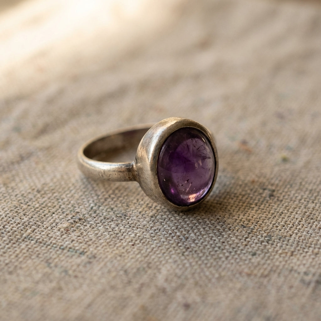 Close up of an ancient amethyst ring in a silver bezel on neutral linen showing patina and deep purple stone under soft directional warm light