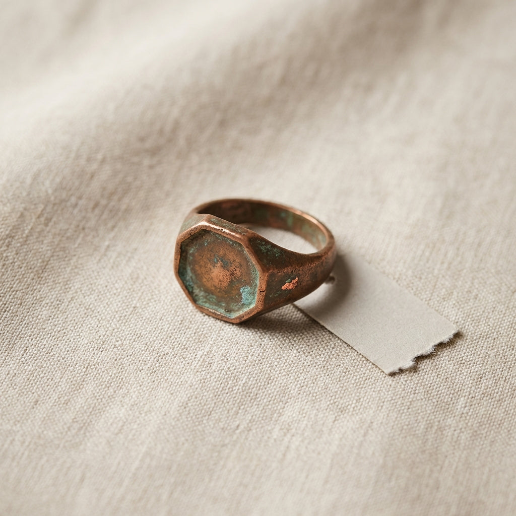 Close textured study of an ancient copper ring octagonal bronze bezel on neutral linen with a blank conservation tag, warm patina and minimalist composition