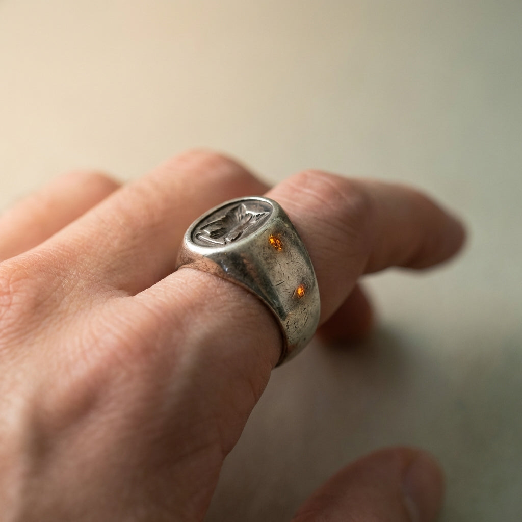 Close up of a hand wearing a worn silver ancient mens ring with carved intaglio showing patina and bezel detail on a minimal warm beige background