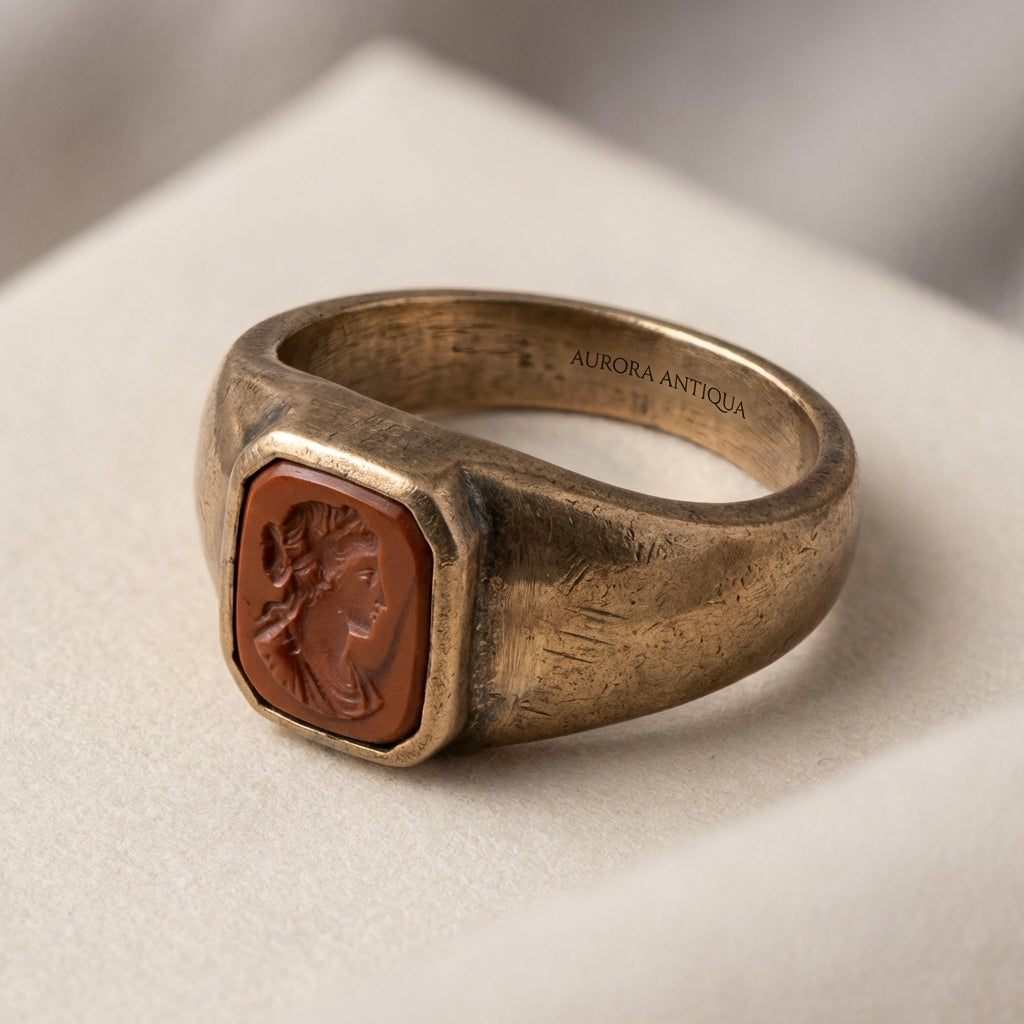 Close up of an ancient signet ring bezel with recessed red jasper intaglio on an aged metal band against a soft beige background in Aurora Antiqua palette