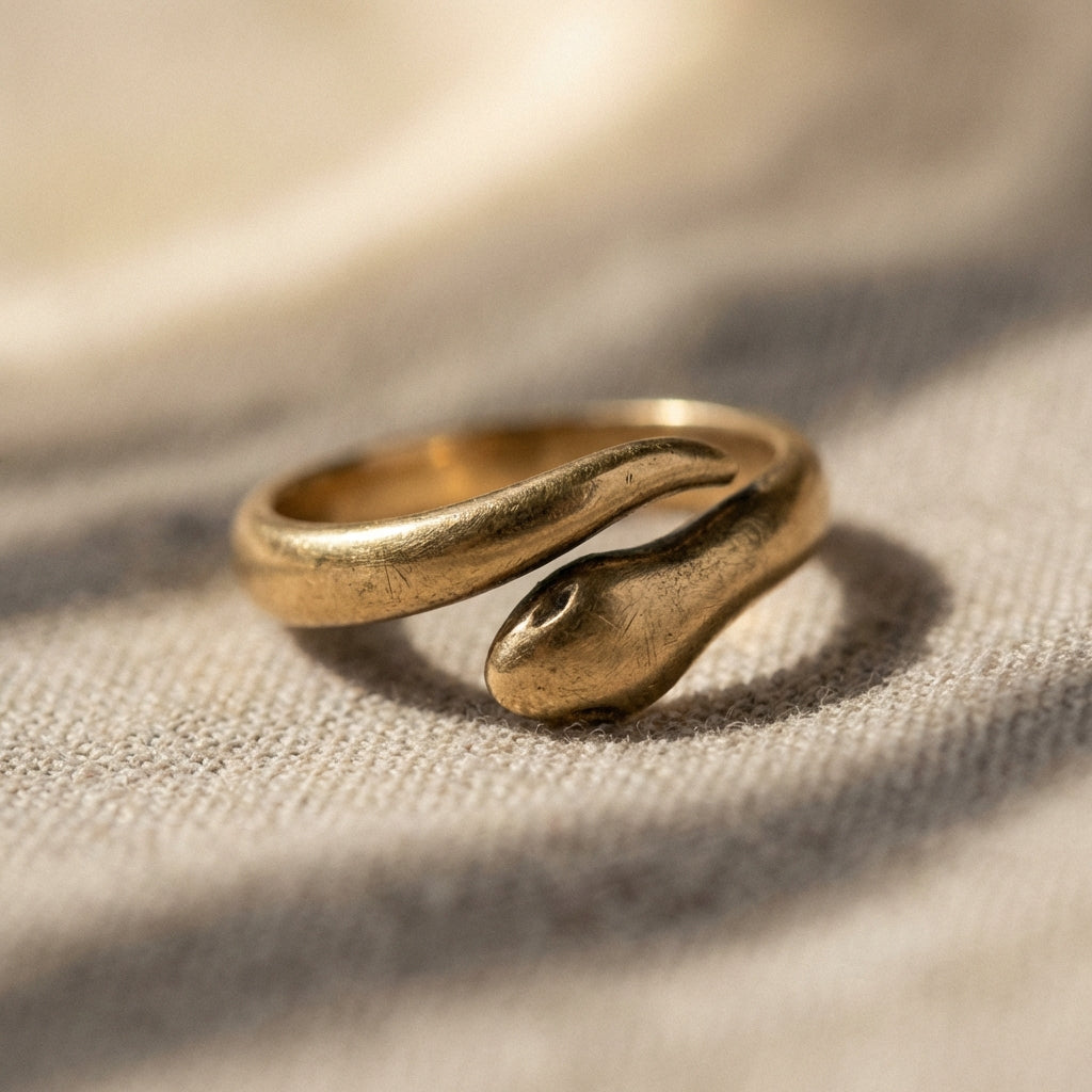 Close photograph of a worn gold ouroboros ring on a neutral textile background ede7da with soft directional warm light revealing texture patina and subtle ae8339 highlights minimal Aurora Antiqua aesthetic
