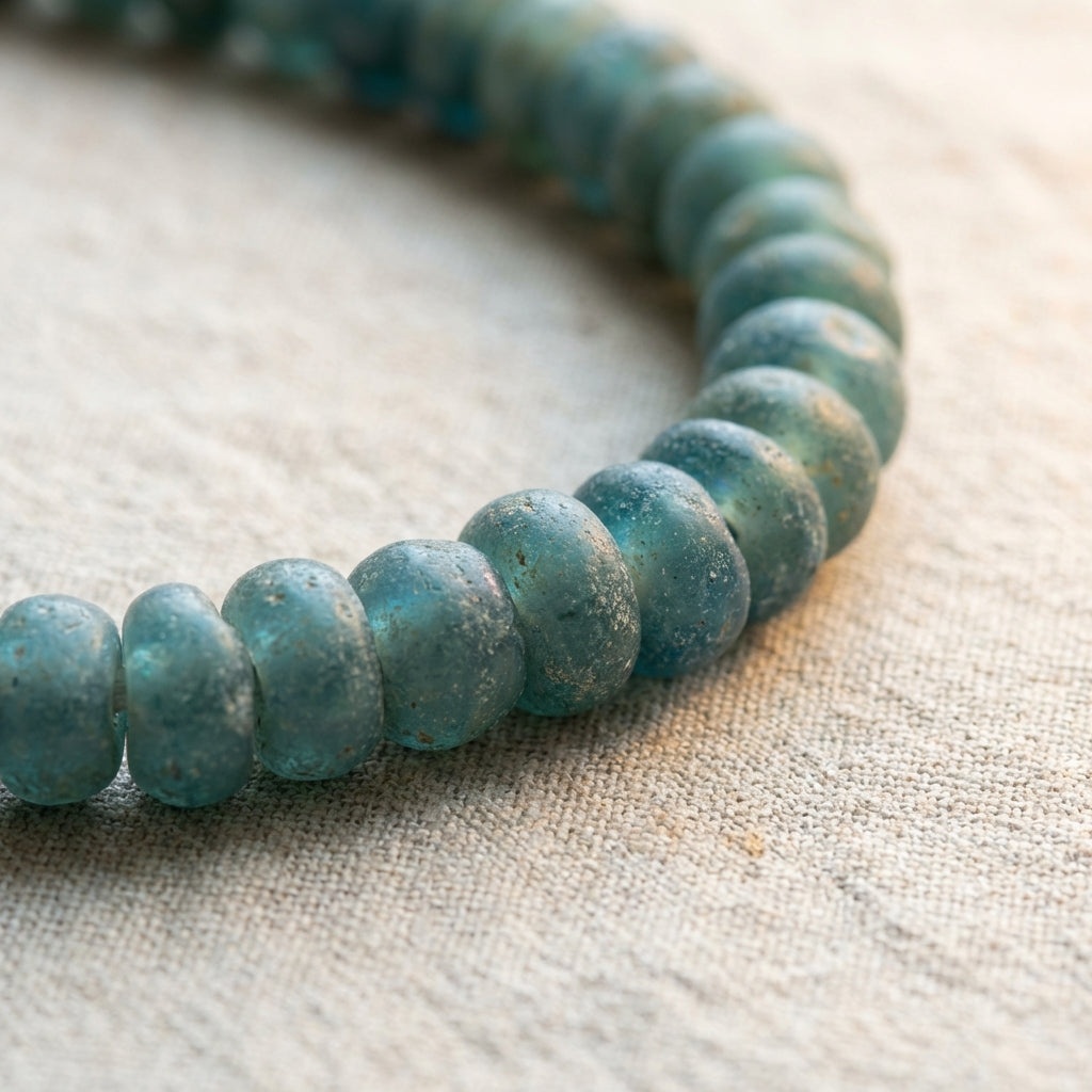 Close up of blue green roman glass jewelry beads on a neutral linen background showing subtle iridescence surface weathering and rounded edges