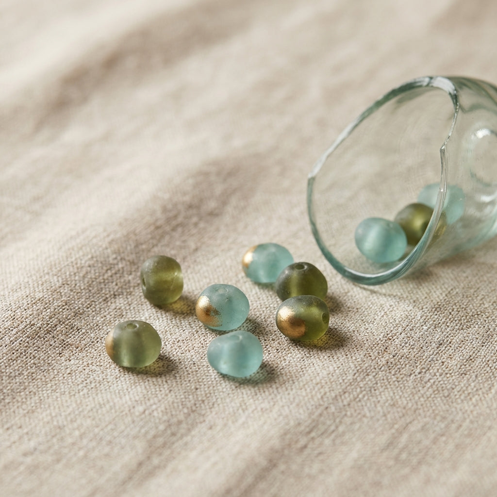 Close focused still life of aqua and olive glass beads and a blown mould glass fragment on neutral linen background showcasing roman glass jewelry in Aurora Antiqua palette